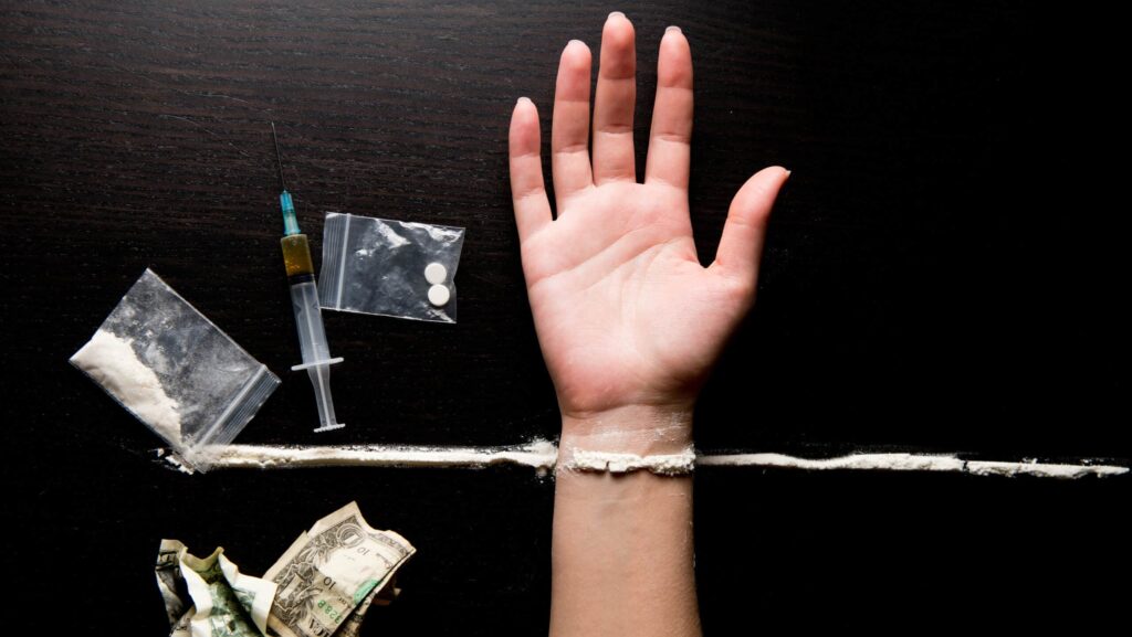 A persons hand is laid out on a dark table next to a syringe, cash, bags of drugs and powdered drugs, which represents meth vs crack similarities and differences.