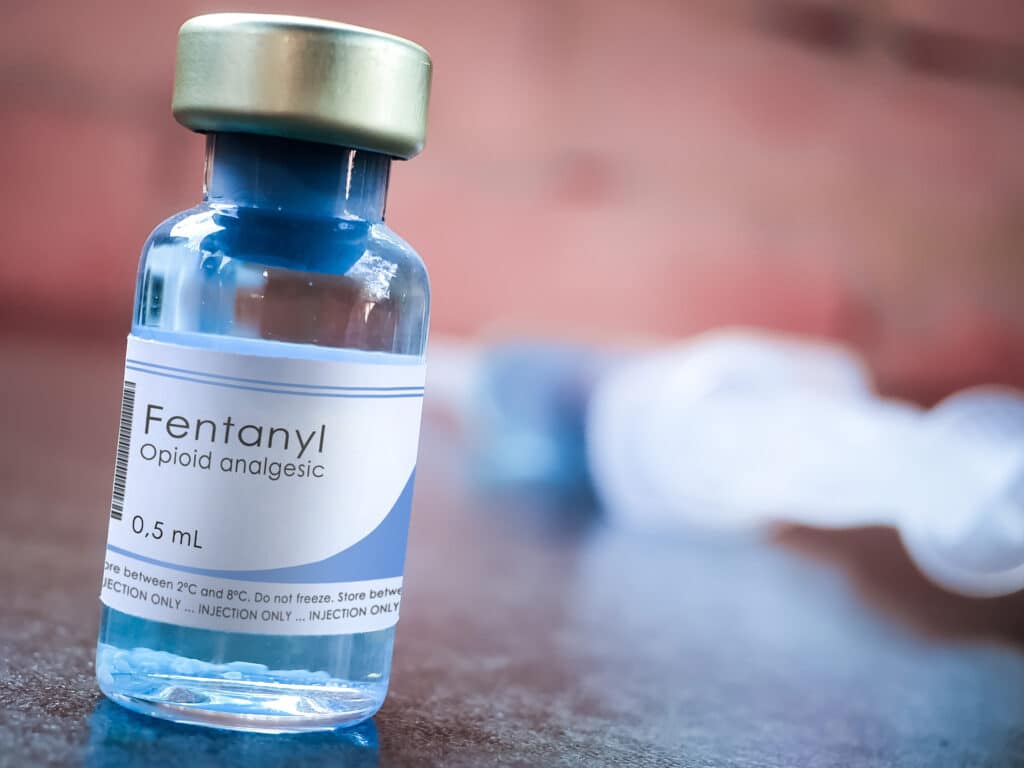 On a blurred red background, a blue-ish glass bottle of liquid fentanyl sits on a table. Users question how long does fentanyl stay in the system?