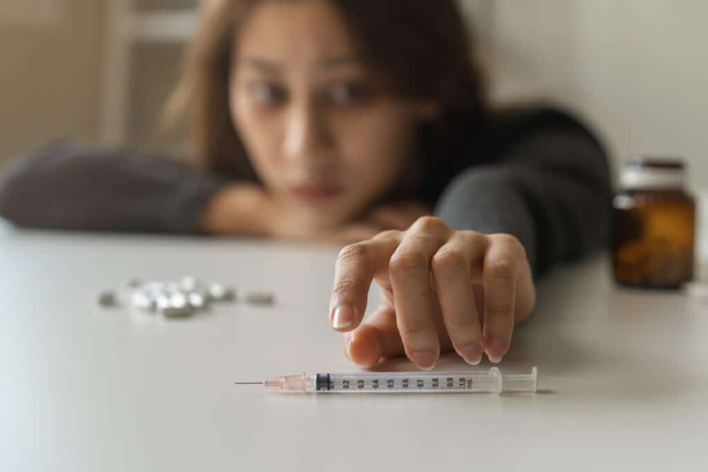 A female young adult taking Xanax and cocaine lays her head on the table with pills while she reaches for a syringe.