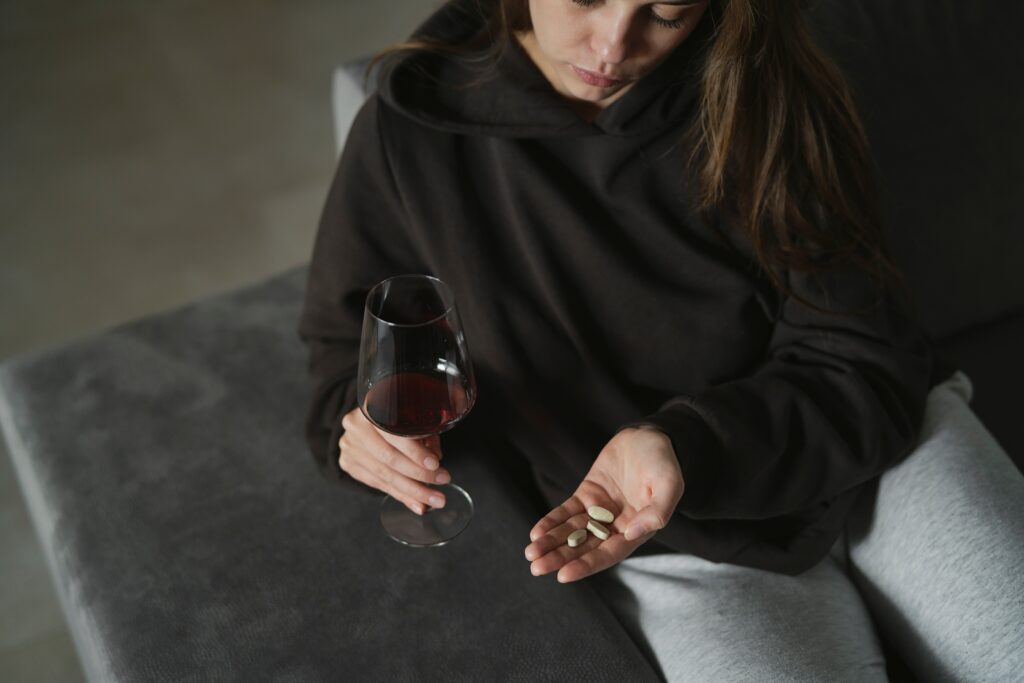 A young women in a dark hoodie is sitting on the sofa holding a glass of wine in one hand and her other hand contains a few pills representing keppra and alcohol.