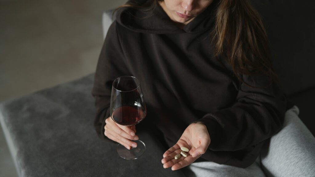 A young women in a dark hoodie is sitting on the sofa holding a glass of wine in one hand and her other hand contains a few pills representing keppra and alcohol.