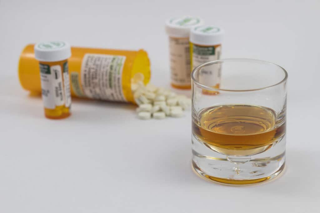 A few prescription bottles and spilled over pills of hydrocodone and alcohol are laid out on a white table.