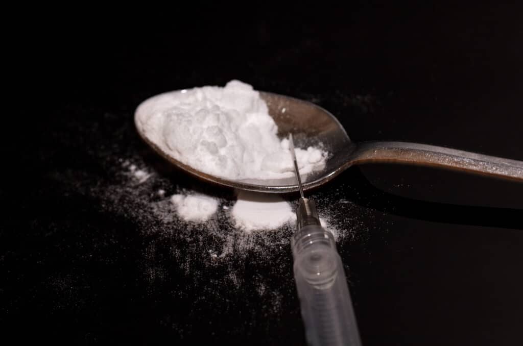 On a black background, a spoon filled with white powder cocaine mixed with heroin lays next to an empty syringe shows what is a speedball.