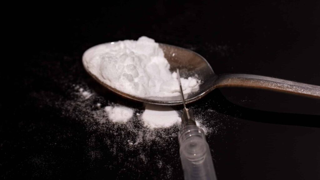 On a black background, a spoon filled with white powder cocaine mixed with heroin lays next to an empty syringe shows what is a speedball.