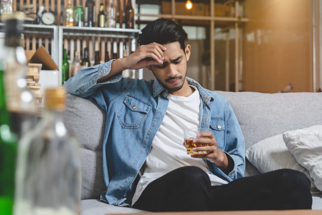 A young man sitting on a sofa holding a glass of alcohol is experiencing side effects of kratom and alcohol use.