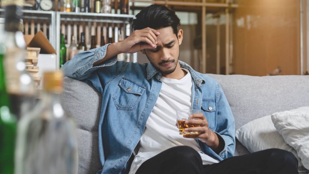 A young man sitting on a sofa holding a glass of alcohol is experiencing side effects of kratom and alcohol use.