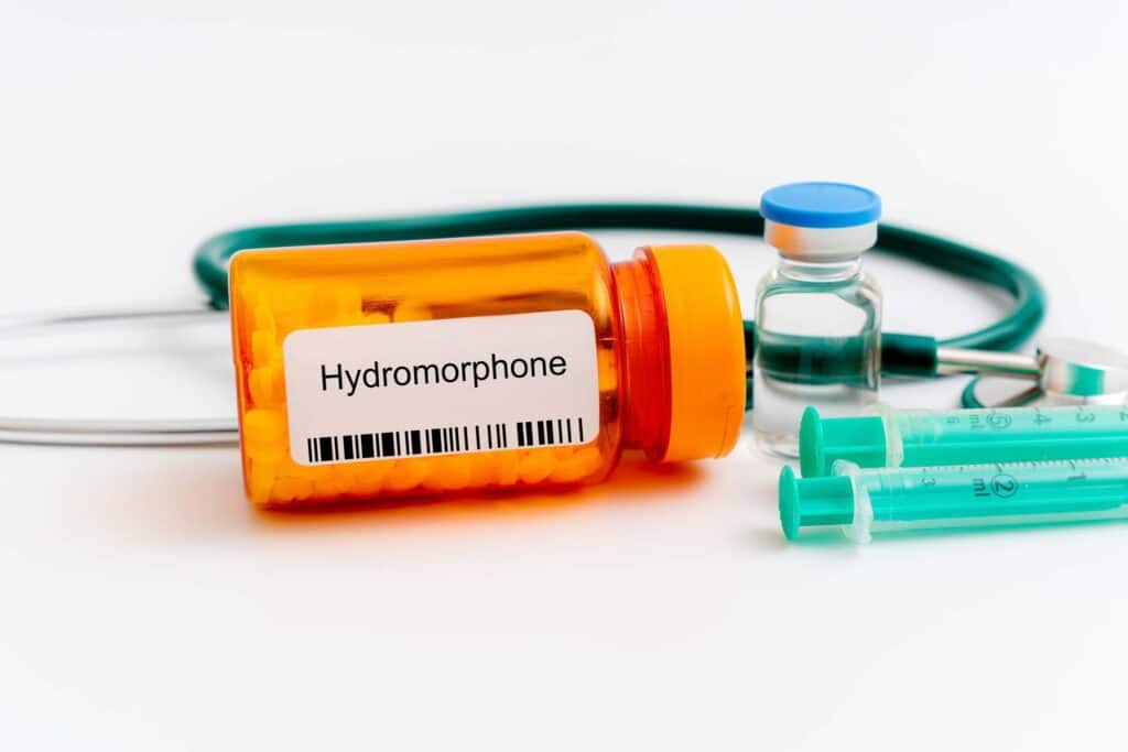 On a white background, an orange bottle of hydromorphone pills is laid out next to a stethoscope, a bottle of clear liquid and syringes. This represents how long Dilaudid stays in the system.