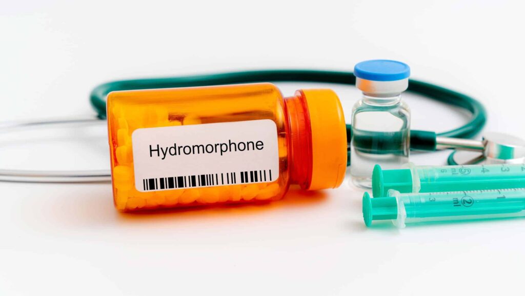 On a white background, an orange bottle of hydromorphone pills is laid out next to a stethoscope, a bottle of clear liquid and syringes. This represents how long Dilaudid stays in the system.