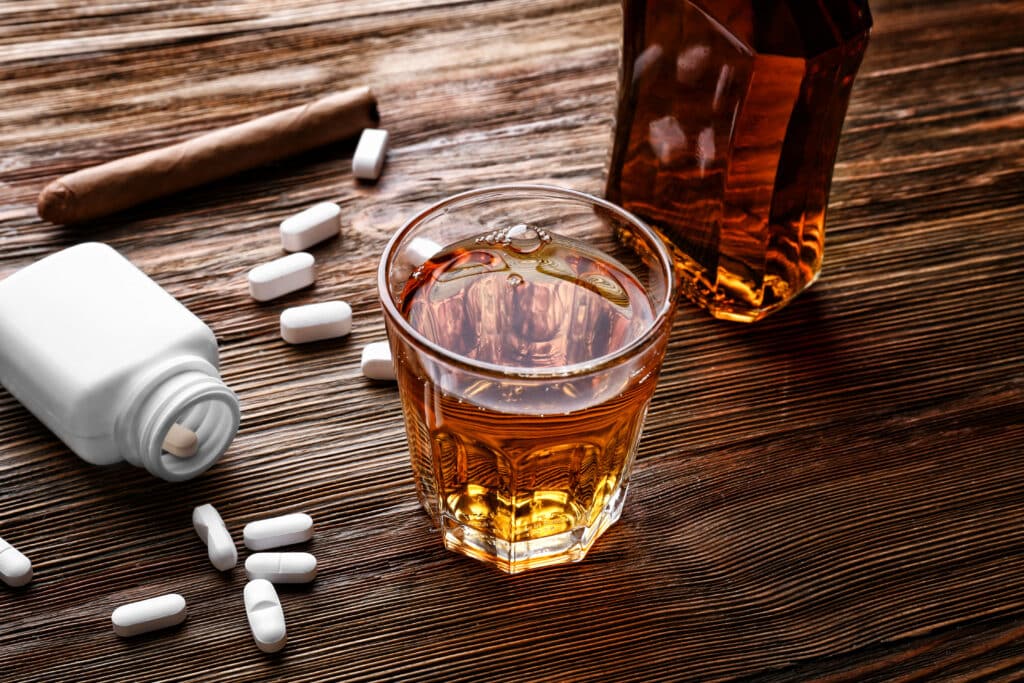 On a wooden table, there is spilled over bottle of klonopin pills next to a glass of alcohol representing the side effects of klonopin and alcohol use.