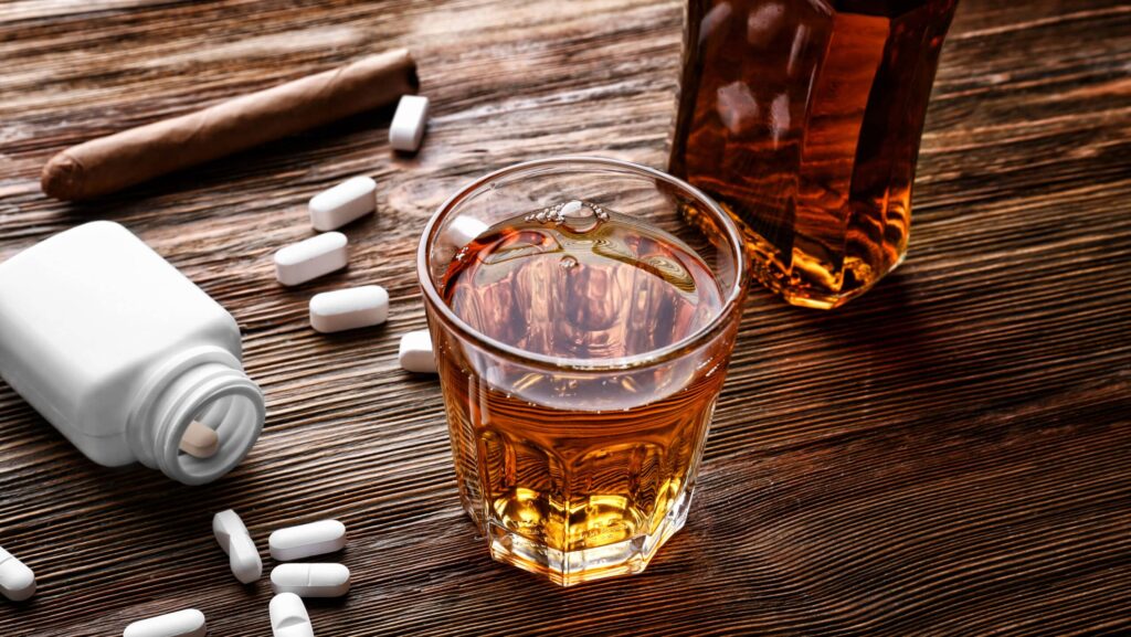 On a wooden table, there is spilled over bottle of klonopin pills next to a glass of alcohol representing the side effects of klonopin and alcohol use.
