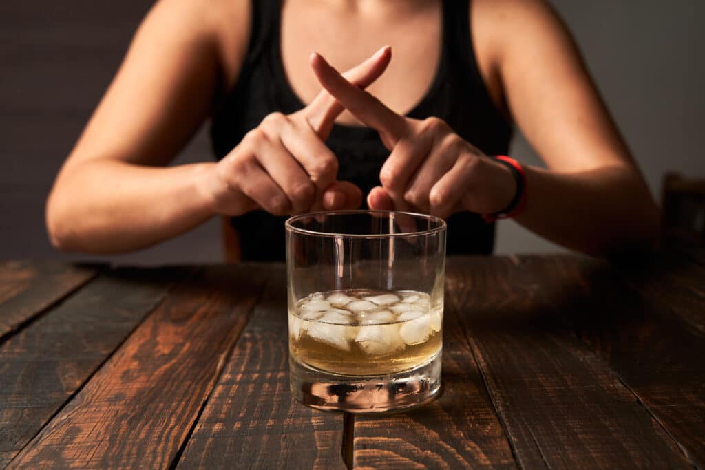 A glass of alcohol sits on a table with a woman behind the glass with her finger crossed in an X form represents the dangers of mixing Claritin and alcohol.