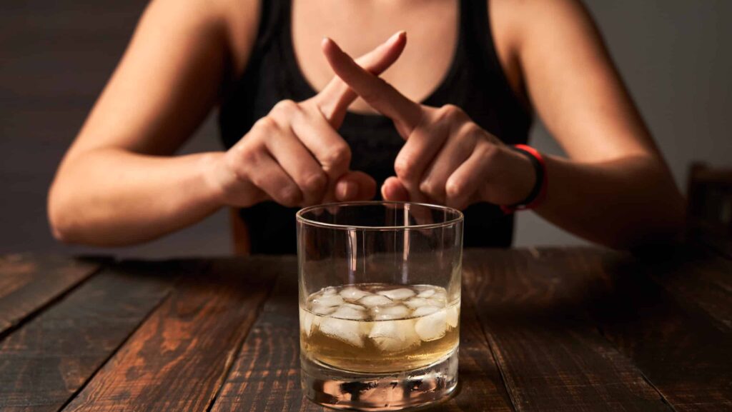 A glass of alcohol sits on a table with a woman behind the glass with her finger crossed in an X form represents the dangers of mixing Claritin and alcohol.