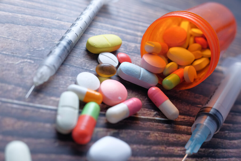 On a wooden table, a spilled over bottle with color pills and tablets lay net to two syringes represents the question of how long does it take to recover from an overdose.