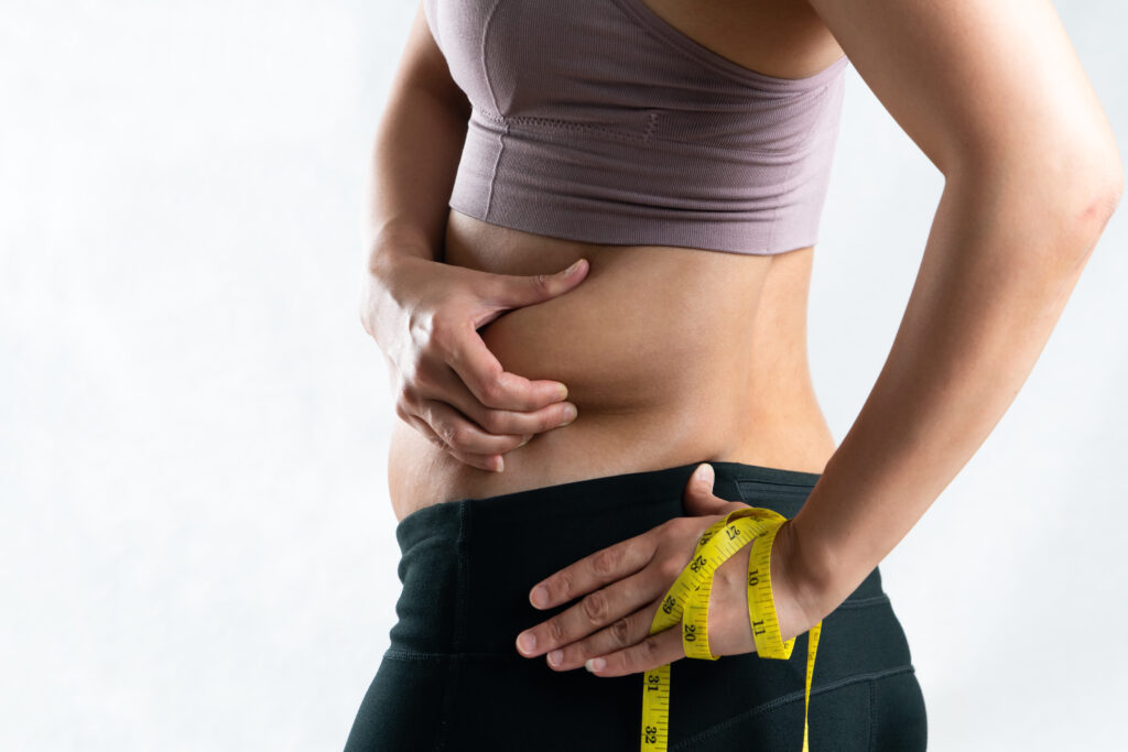 A woman in gym clothing is pinching her side stomach while her other had is holding a measuring tape and representing the possibility of weight gain from buspirone.