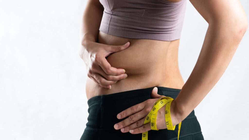 A woman in gym clothing is pinching her side stomach while her other had is holding a measuring tape and representing the possibility of weight gain from buspirone.