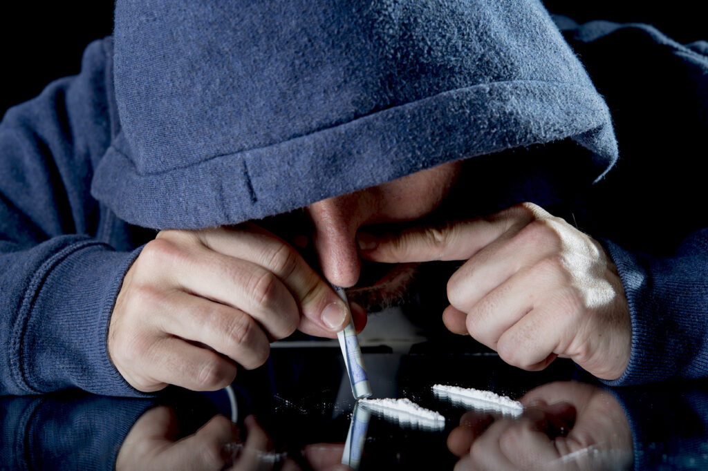A man in a blue hoodie is snorting Gabapentin from a glass table with a rolled up dollar bill.