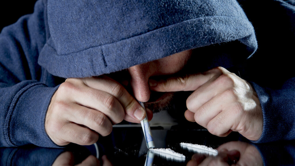 A man in a blue hoodie is snorting Gabapentin from a glass table with a rolled up dollar bill.