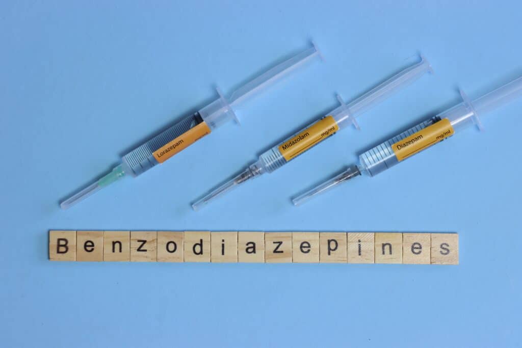 On a blue background there are three syringes of benzos laying next to the word benzodiazepines spelled out in little square letters. This represents different types of benzos medications such as Ativan.
