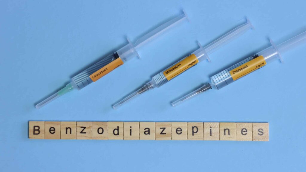 On a blue background there are three syringes of benzos laying next to the word benzodiazepines spelled out in little square letters. This represents different types of benzos medications such as Ativan.