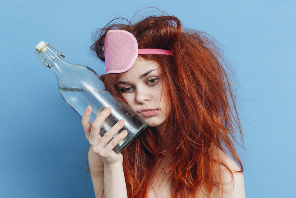 A red headed young girl is wearing a pink sleeping mask on her head while holding a glass bottle of water against her cheek represents how long it takes to try and sober up.