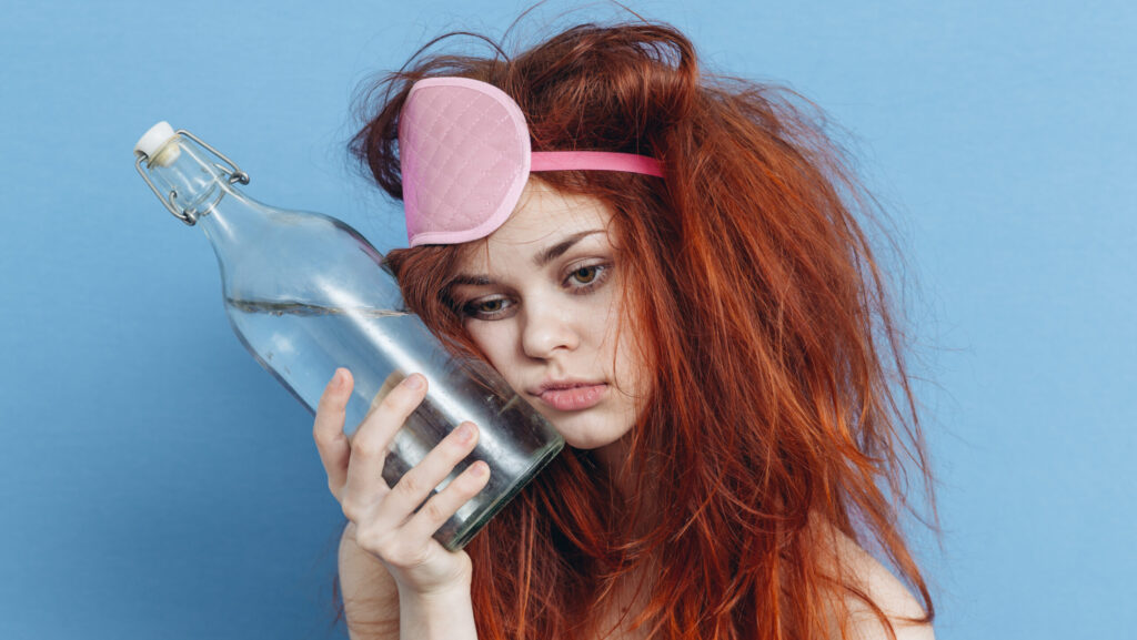 A red headed young girl is wearing a pink sleeping mask on her head while holding a glass bottle of water against her cheek represents how long it takes to try and sober up.