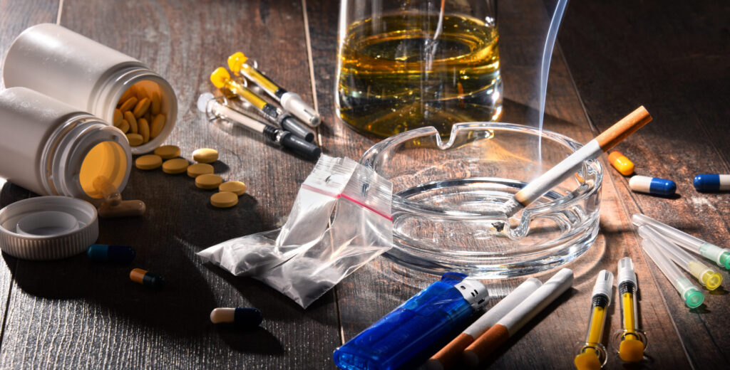 On a table, there is a glass of alcohol, opioid pill bottles will pills spilling out, syringes and cigarettes laying next to each other representing the most dangerous drugs.