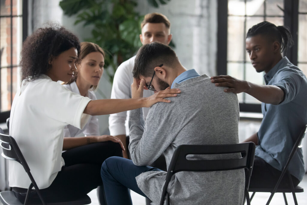 A group of young diverse adults sit in a circle during a SMART recovery meeting and console a young man in glasses.