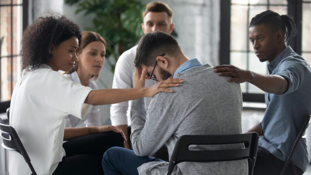 A group of young diverse adults sit in a circle during a SMART recovery meeting and console a young man in glasses.