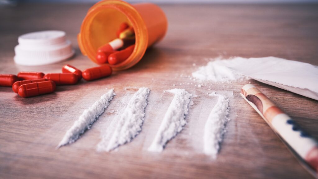 On a table, there is a spilled over bottle of red pills, four lines of cocaine and a rolled up bill. People often question whether or not cocaine is a depressant or stimulant.