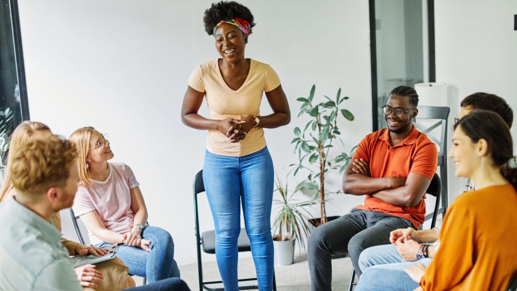 A group of diverse young adults sit in a circle during a PHP or IOP group session while one young women stands to speak about her addiction.