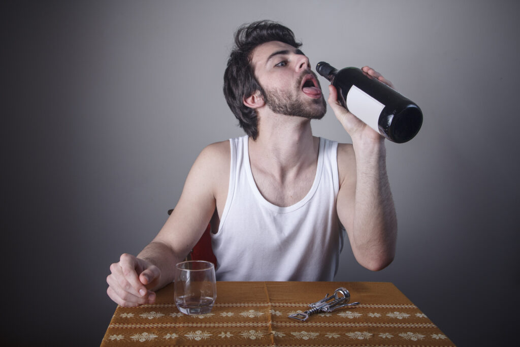 A young man is sitting at the table attempting to drink from a full bottle of wine represents a person who might have dry drunk syndrome.