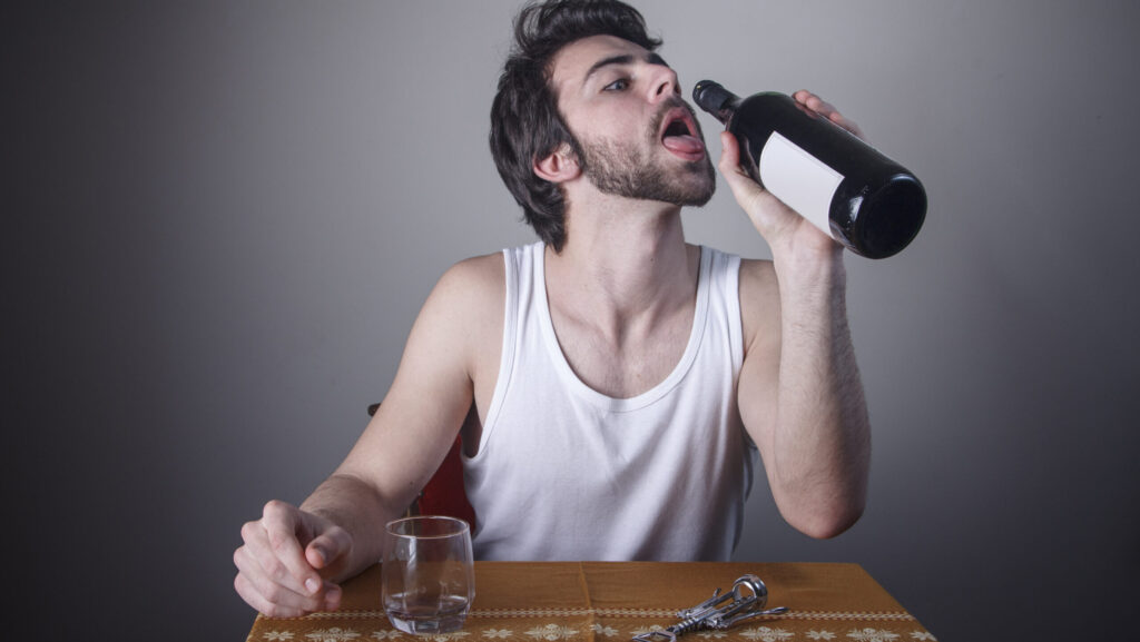 A young man is sitting at the table attempting to drink from a full bottle of wine represents a person who might have dry drunk syndrome.