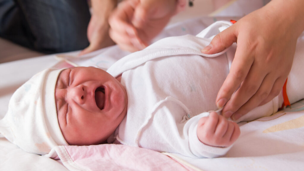 A newborn baby white a white hat and onesie is crying because it is going through cocaine withdrawals.