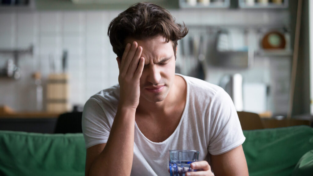 A young man in a white shirt is holding a cup of water with his hands on his temples because he is experiencing a headache from Seroquel withdrawal.