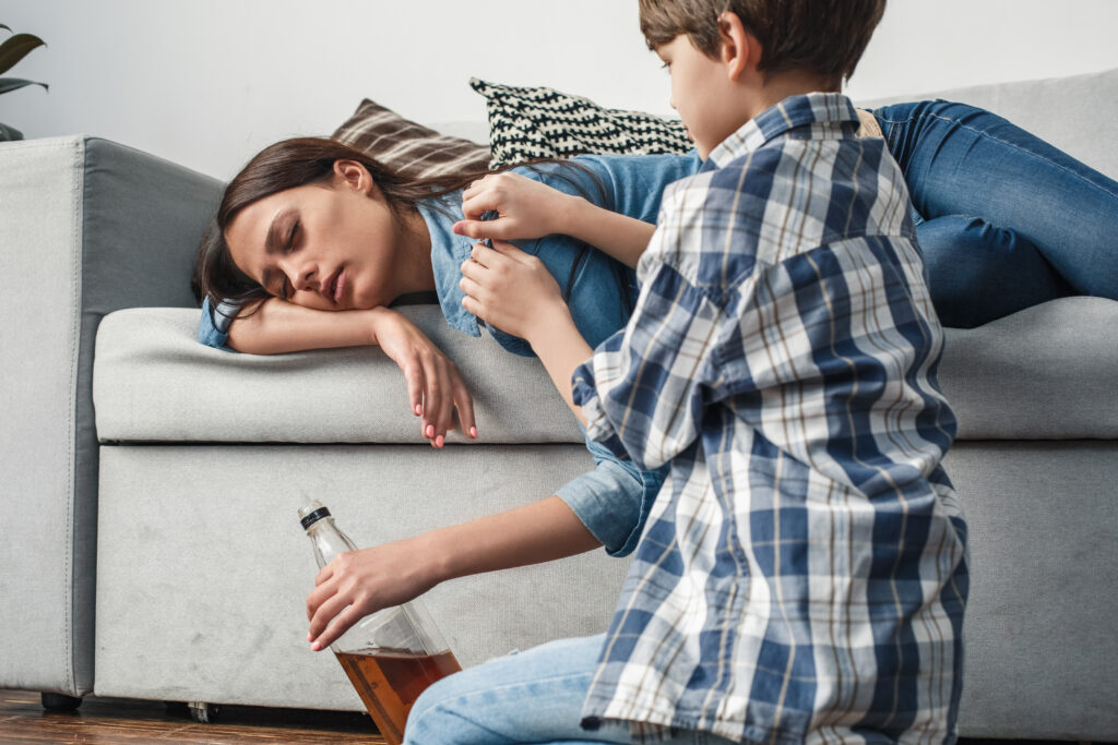 A young boy confronting his mother as she lays on sofa passed out with a bottle of alcohol in her hand.
