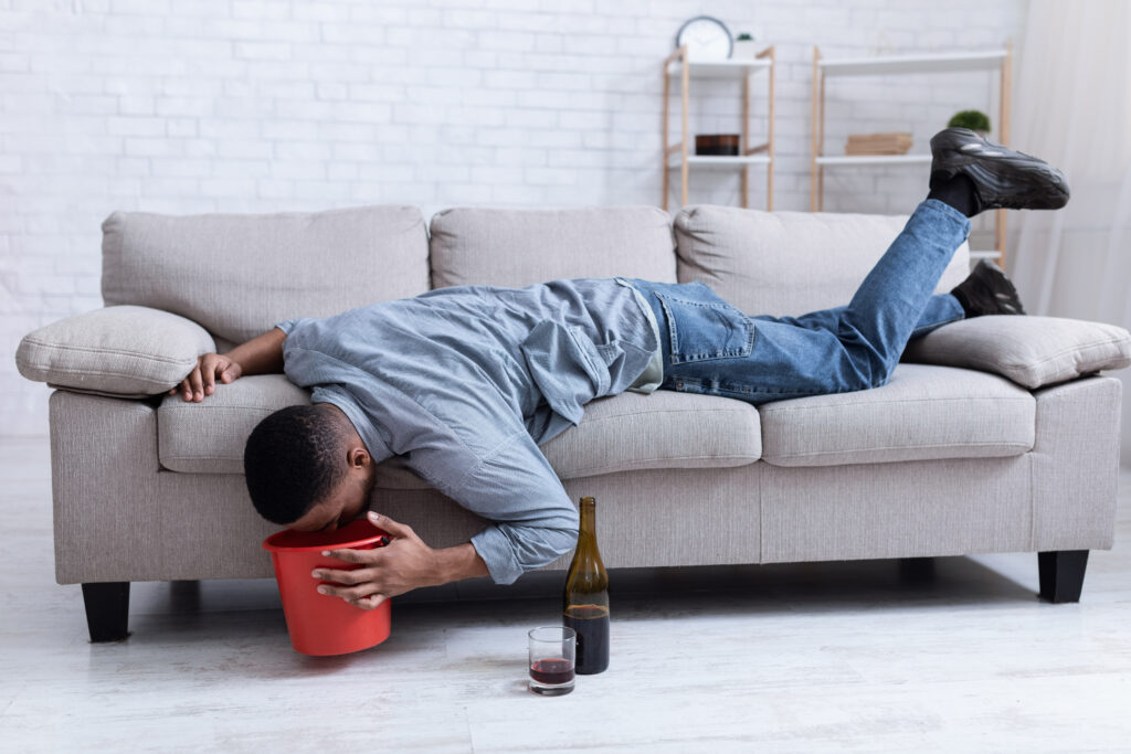 A man laying on the sofa with his head over a bucket next to a bottle of wine is experiencing alcohol poisoning.
