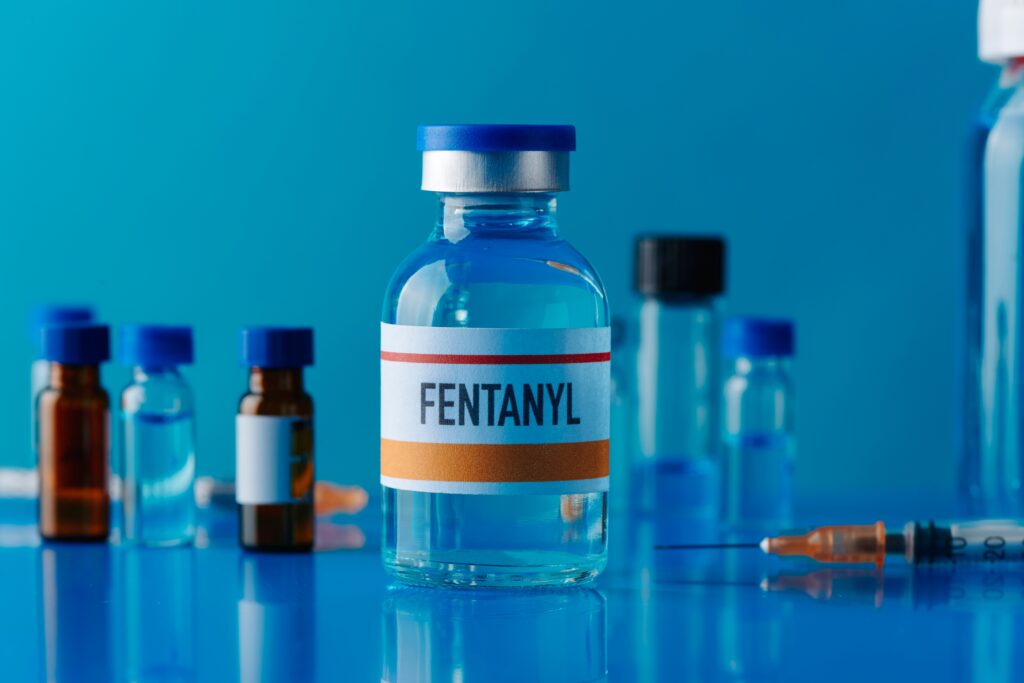 A glass bottle of fentanyl sits on a table with other glass vials and a syringe in the background.