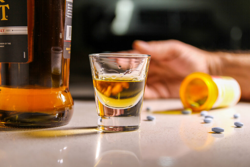 A shot glass of alcohol next to a spill over pill bottle of morphine represents the dangers of mixing morphine and alcohol.