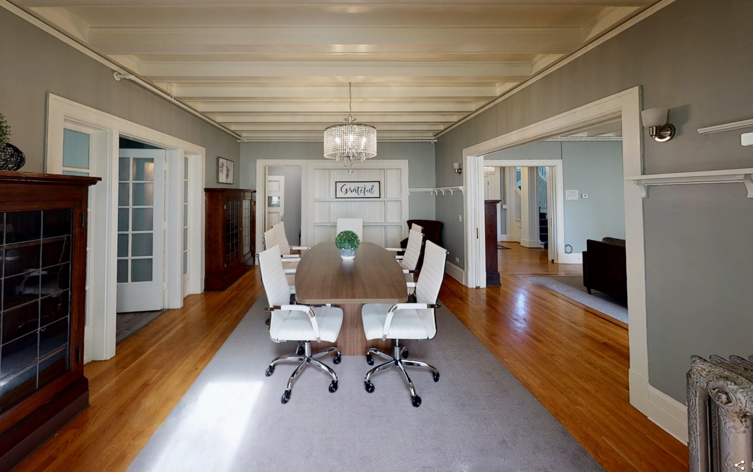 interior of dining room, with white molding, crystal chandelier and a wood table with 6 office chairs and sign that reads "Grateful"
