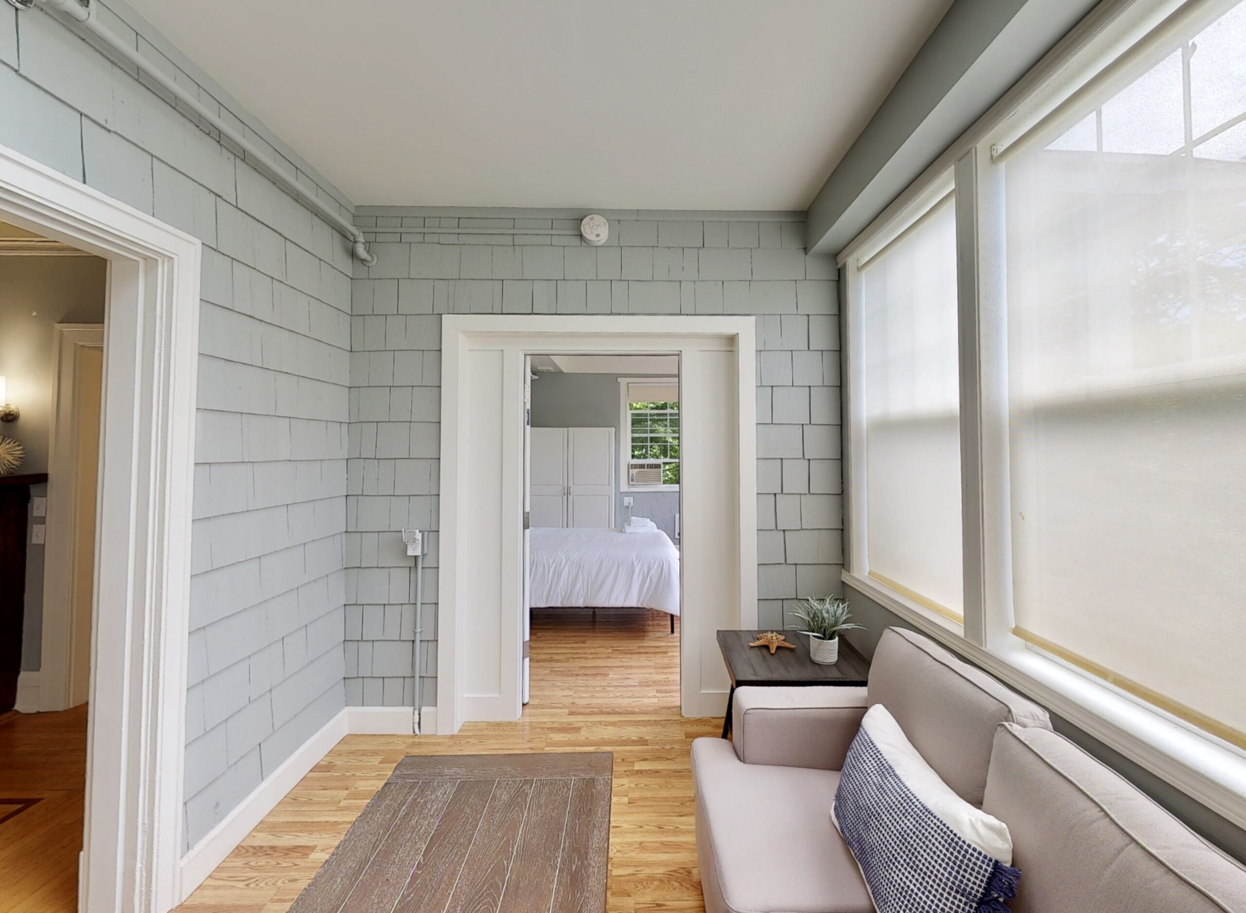 Sitting area with grey couch, large windows, wood floors and door opening to clean, airy bedroom with a bed that has white bedding and white cabinet