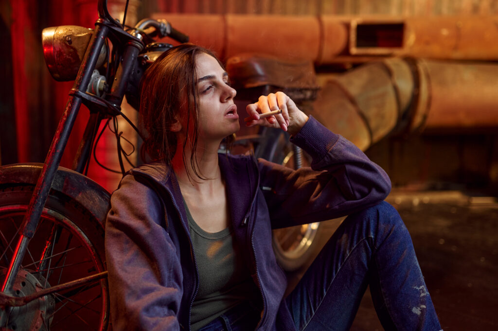 A disheveled looking woman leaning on the floor and smoking a cigarette is a sign of how to tell if someone is on drugs.