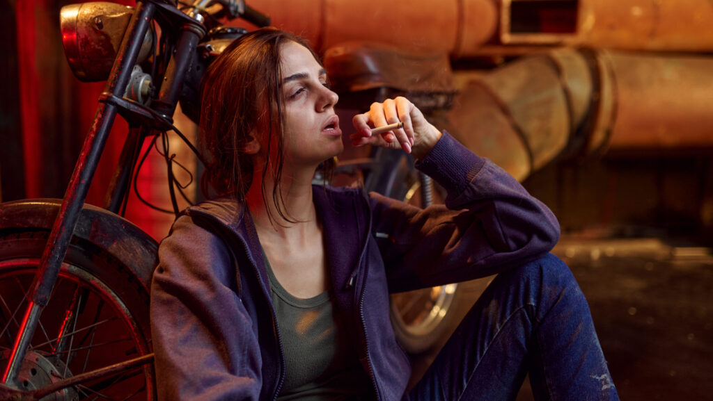 A disheveled looking woman leaning on the floor and smoking a cigarette is a sign of how to tell if someone is on drugs.