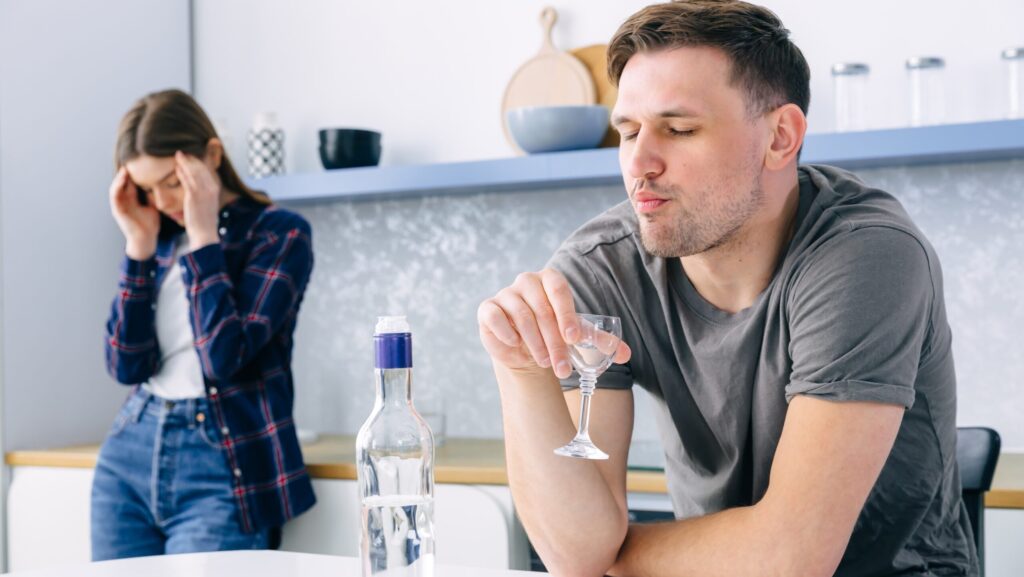 A woman rubbing her temples at her male friend who is an alcoholic and is drinking.