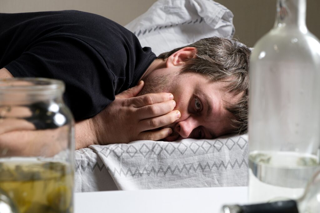 A man laying on a sofa, looking disheveled and visibly experiencing DTS after quitting the alcoholic drinks that are surrounding him.