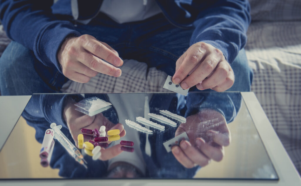 A man habitually using drugs doing a line of cocaine next to pills and syringes on a table.