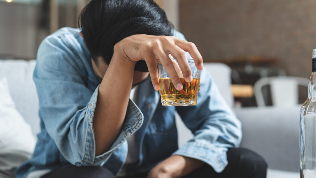 A man with his head down holding a glass of alcohol is drunk and wondering how long he will stay drunk.