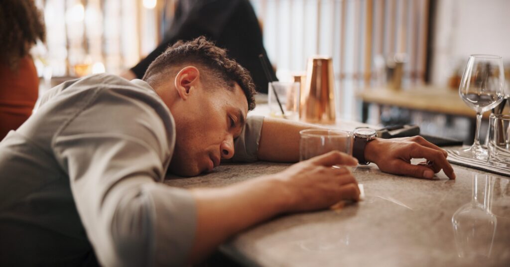 A man slumped over the counter holding his drink is experiencing alcohol fatigue.
