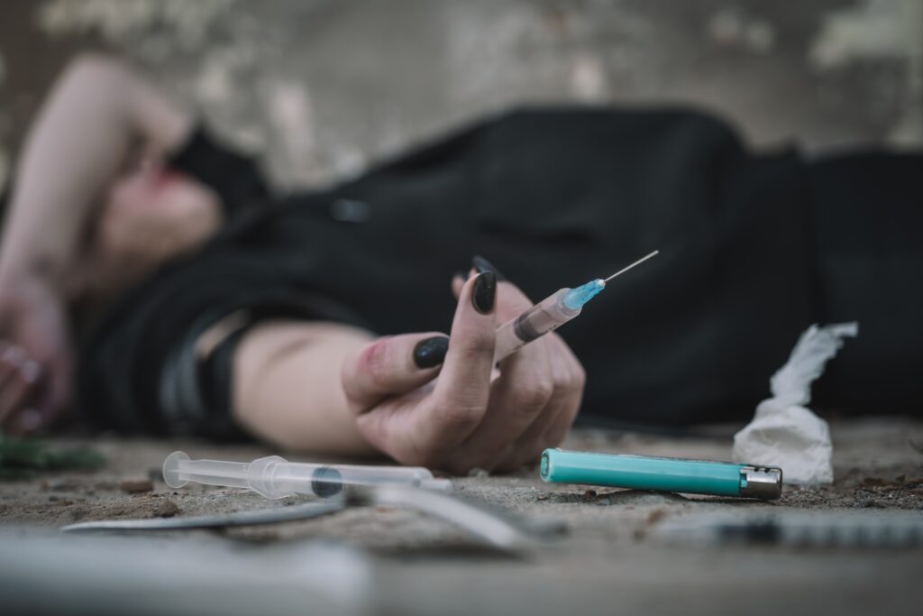 A woman is laying on the ground holding a drug syringe contemplating using a shared needle.