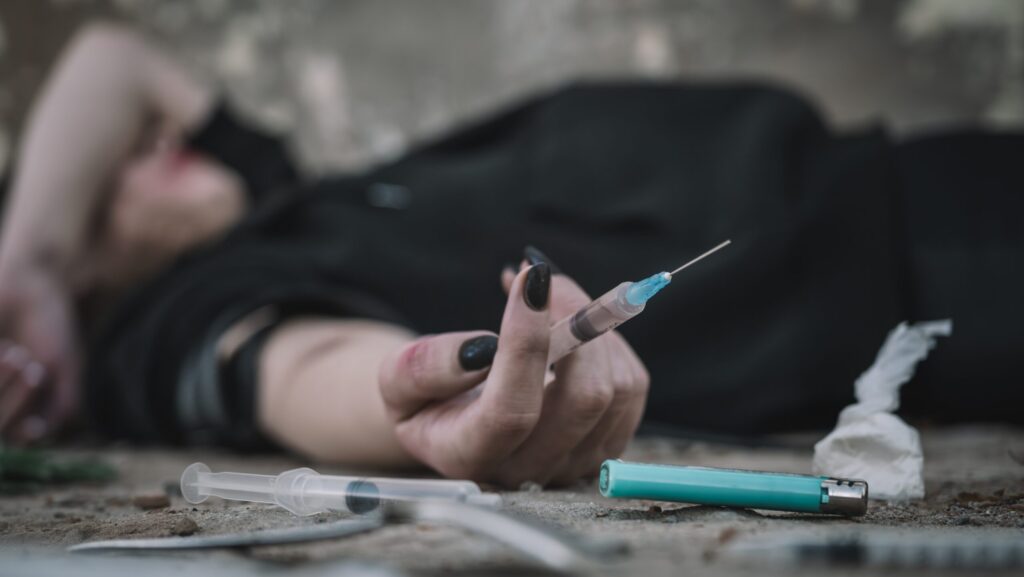 A woman is laying on the ground holding a drug syringe contemplating using a shared needle.