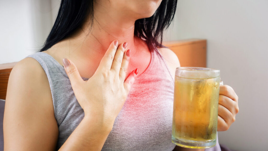 A young women holding a glass of beer with her hand on her chest because she is wondering can alcohol cause stomach problems like acid reflux.
